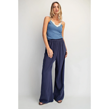 Woman wearing a blue tank top and loose navy pants posing against a white wall.