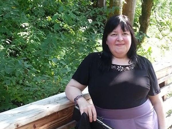 Woman standing outdoors by a wooden railing with trees in the background.