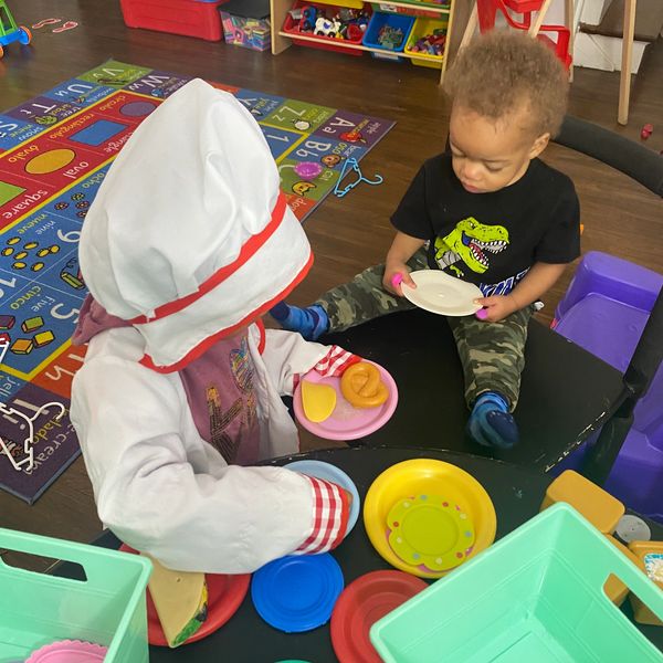 Two toddlers playing pretend at a small table with toy food and dishes.