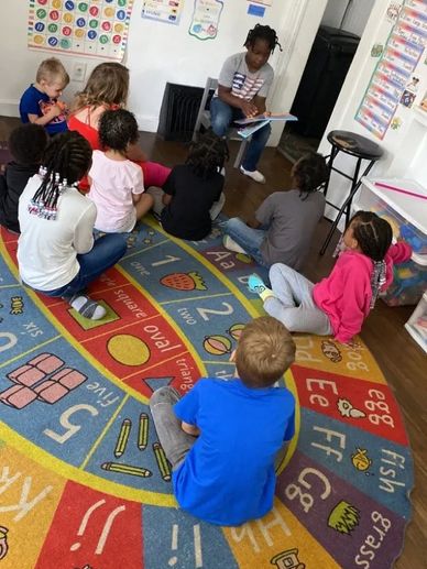 A teacher reads to children sitting on a colorful educational rug.