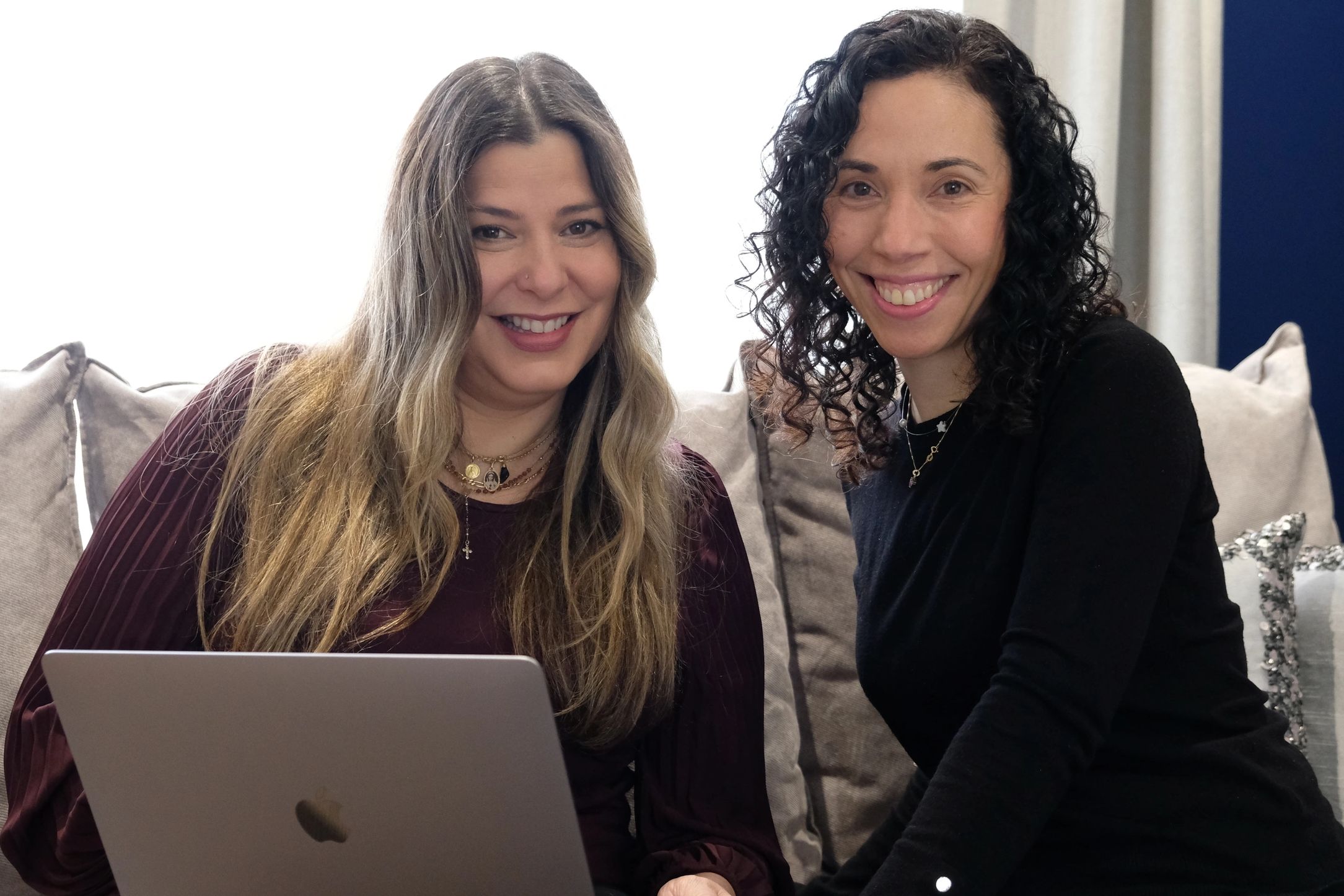 Two women smiling while using a laptop on a couch.