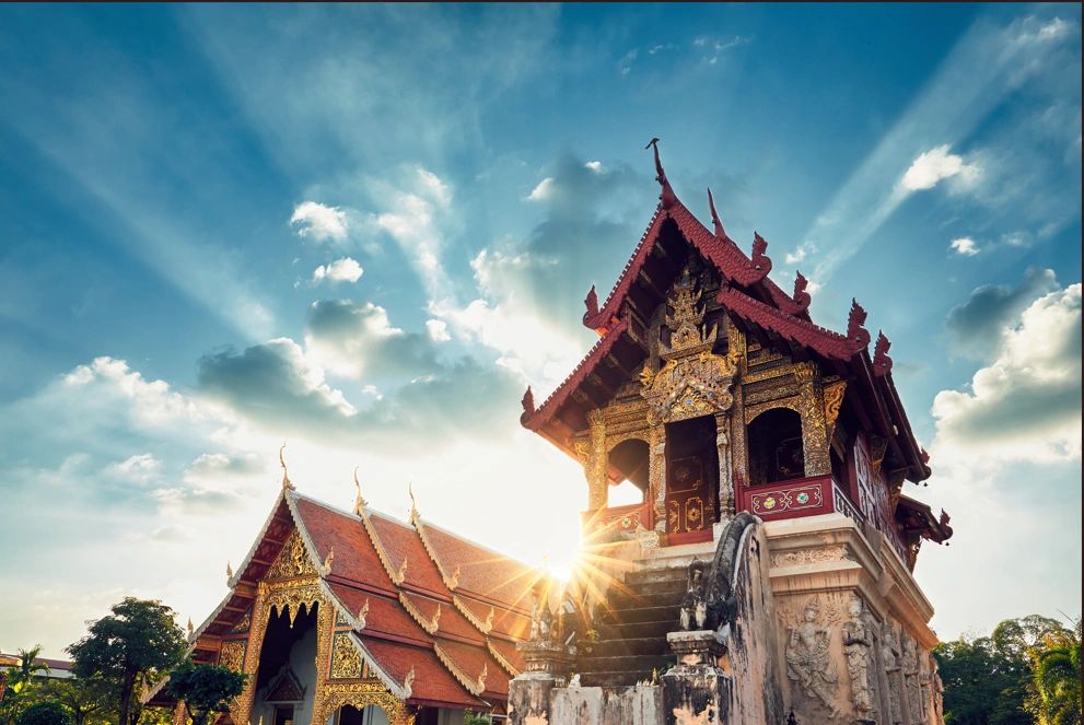 Sunlight shines behind traditional Thai temple buildings under a blue sky.