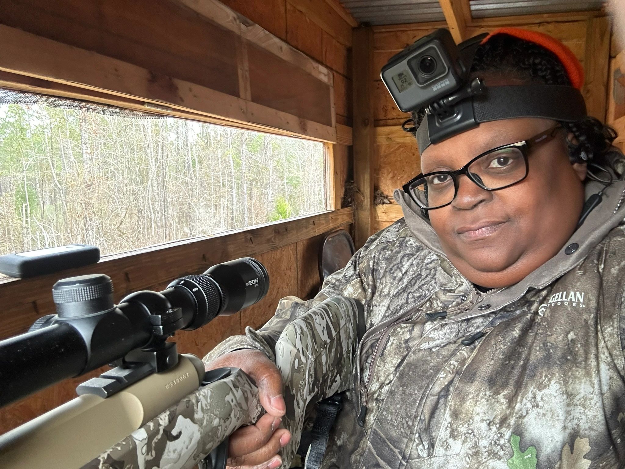 Person in camouflage holding a scoped rifle inside a wooden blind