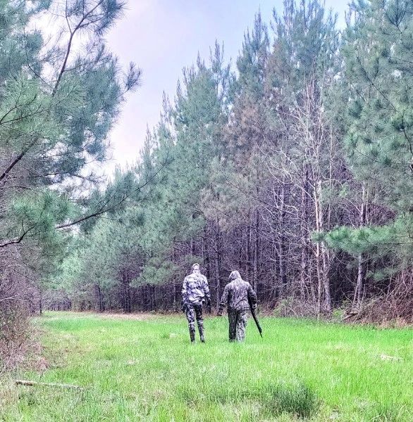 Two people walking in a forest with one carrying a shotgun.