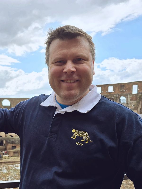 Smiling man in a navy blue shirt with a leopard emblem at an ancient ruin site.