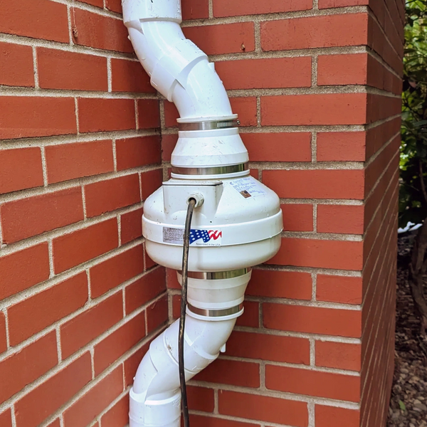 Exterior radon fan mounted on a brick wall with white PVC piping running to and from the fan.
