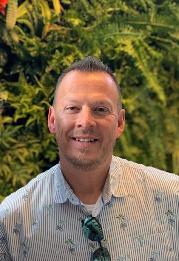 Smiling man in a striped shirt with sunglasses, standing against lush green foliage.