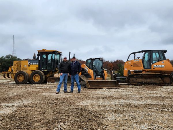 Two men stand in front of heavy construction equipment on a cloudy day.