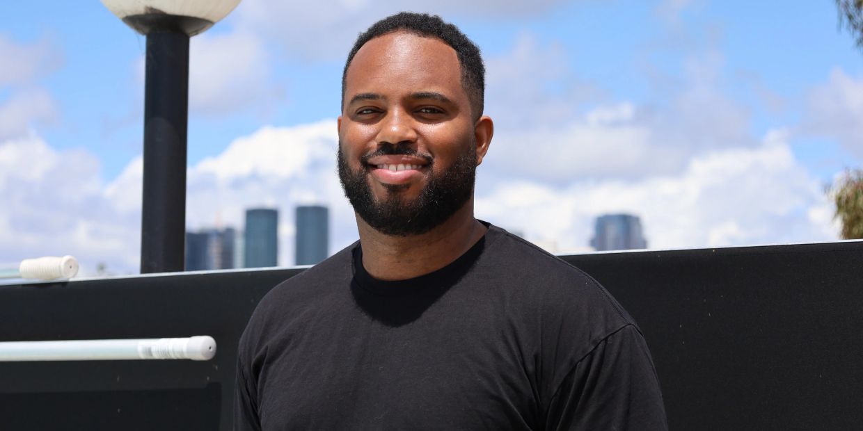Smiling man with a beard wearing a black shirt outdoors against a city skyline.