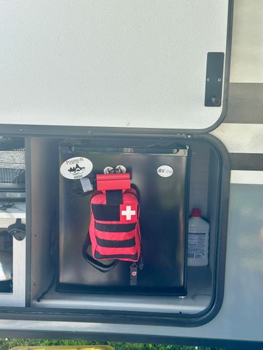 Compact black mini fridge with a red and black first aid kit attached inside a camper storage compartment.