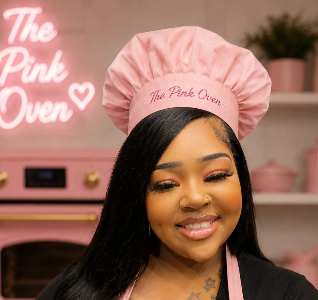 Smiling woman wearing a pink chef hat and apron in a pastel-themed kitchen.