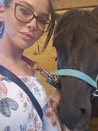 Woman with glasses taking a selfie with a black horse in a stable.