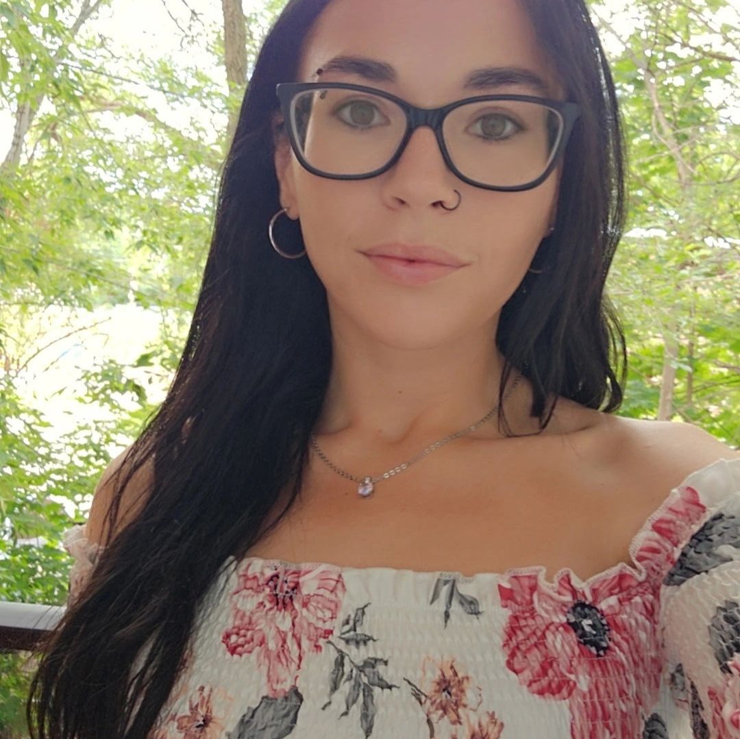 Woman with glasses and floral dress smiling outdoors.