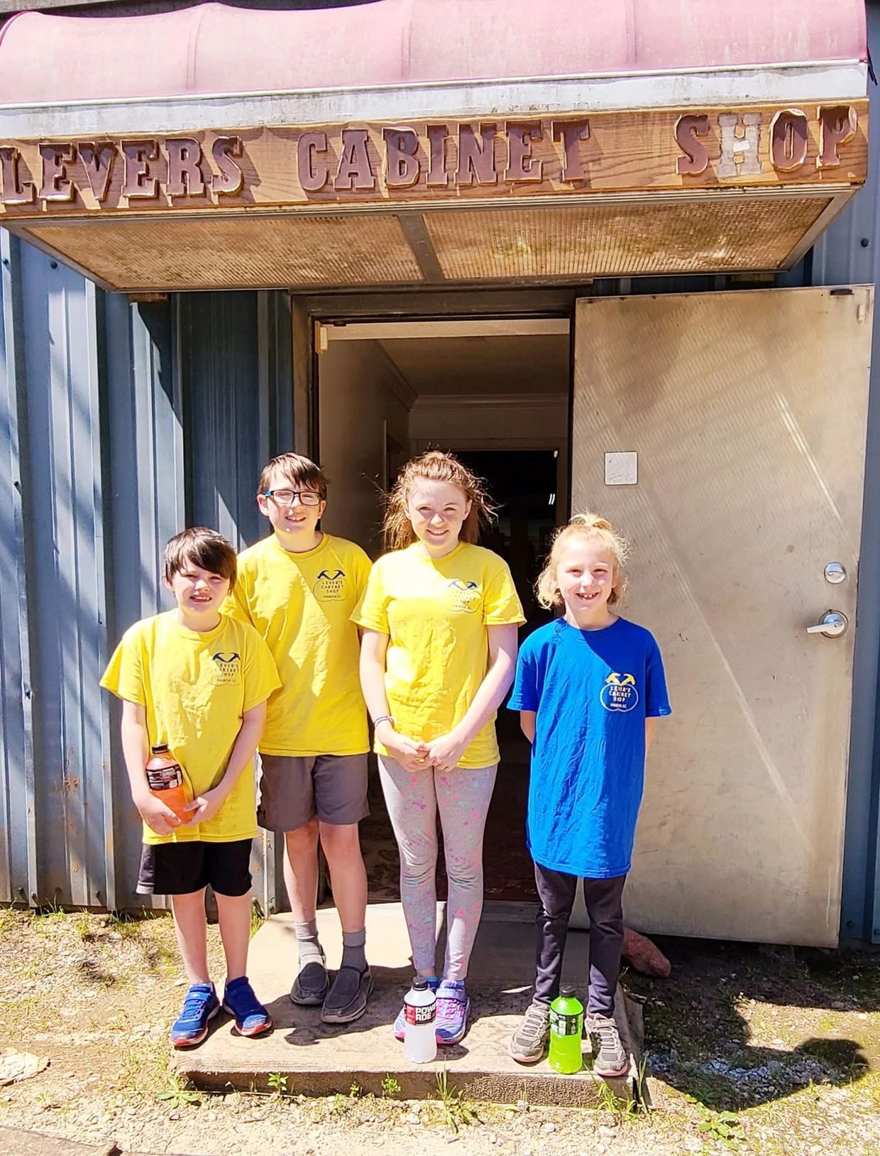 Four children in colorful t-shirts stand outside a cabinet shop on a sunny day.