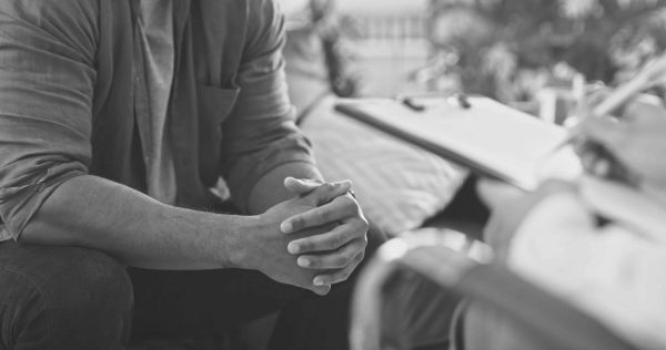 A person sits with hands clasped, speaking to a professional with a clipboard.