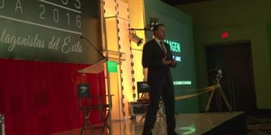 A man in a suit speaks on stage at a formal event with a red and green backdrop.