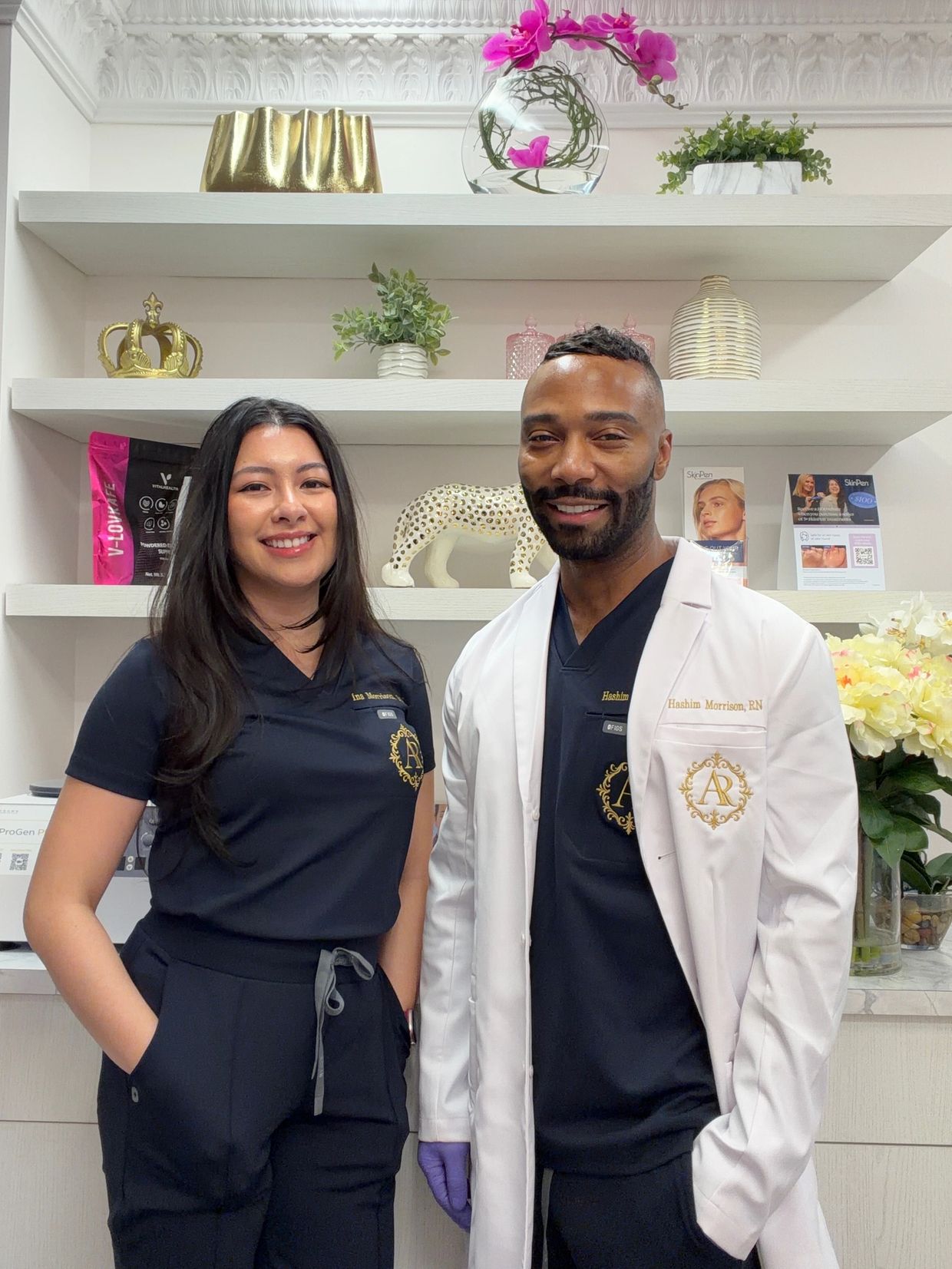Two medical professionals smiling in a clinic with decorative shelves behind them.