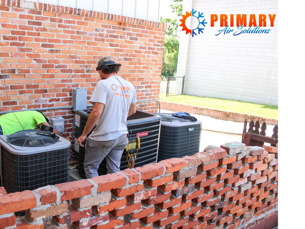 Two technicians servicing outdoor HVAC units beside a brick wall.