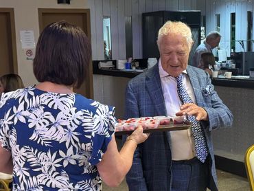 An elderly man in a suit receiving a tray of desserts from a woman.