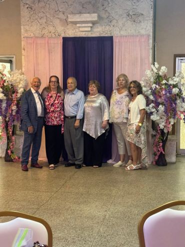 A group of six adults posing in front of pink and purple drapes with floral arrangements.