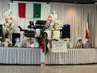 Woman speaking at an Italian-themed event with flags and decorations.