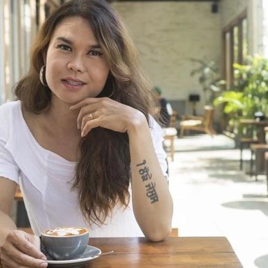 asian trans female in a white top, brunette, tattoo on her arm drinking a coffee sitting at a cafe 