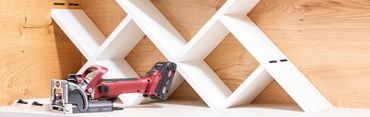 A red power drill resting on a wooden surface next to a white geometric shelf.
