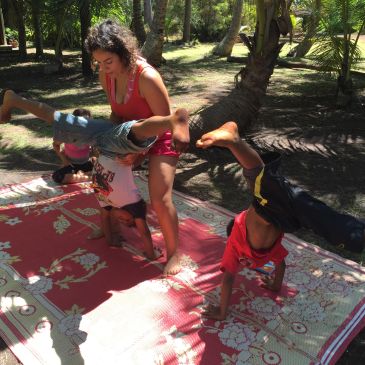 Two children doing handstands with an adult assisting one on a patterned mat outdoors.