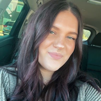 Woman smiling inside a car with dark hair and facial piercings.