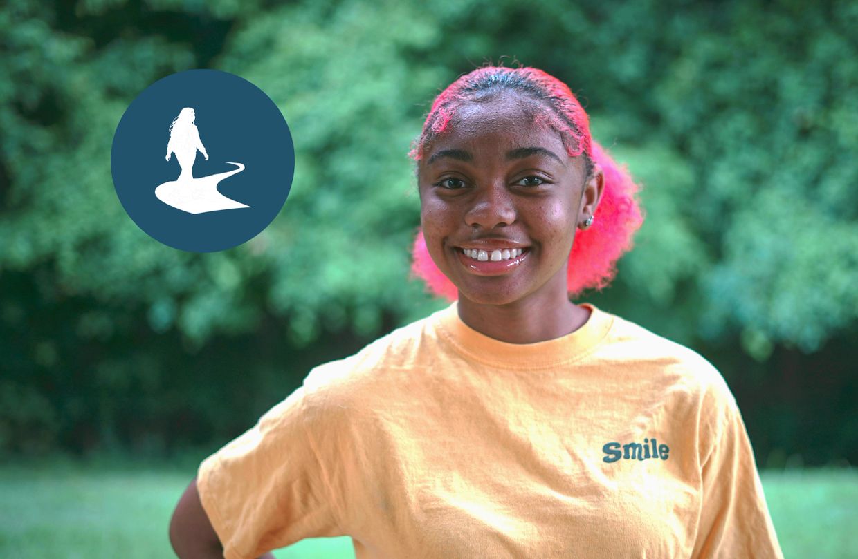 Smiling young woman with pink hair outdoors, wearing a yellow shirt that says Smile. Her Path Forward