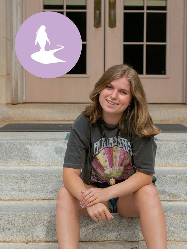 Young woman sitting on stone steps, smiling, wearing a dark graphic t-shirt. Her Path Forward