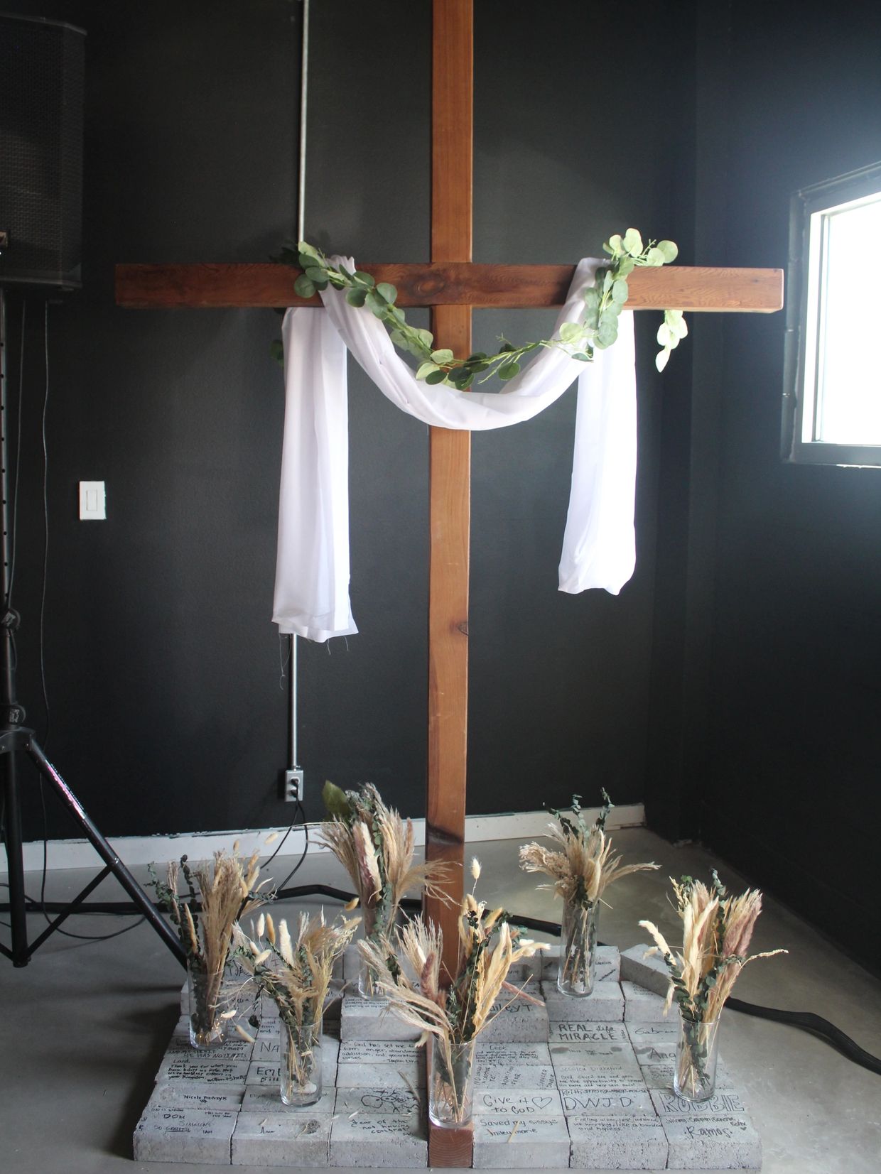 Wooden cross decorated with white cloth and greenery, surrounded by vases of dried flowers on a brick platform.