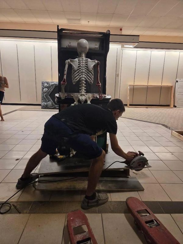 Person using a saw near a seated skeleton model.