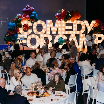 People attending a Comedy For Camp event with illuminated sign and decorations.