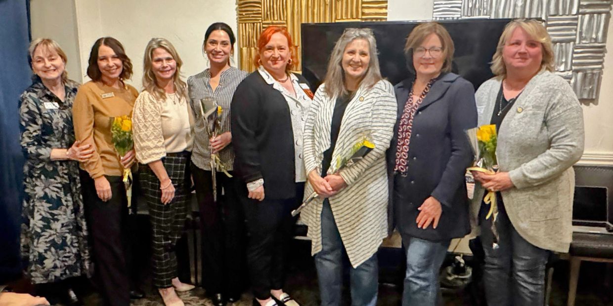 Group of nine women posing indoors with flowers.
