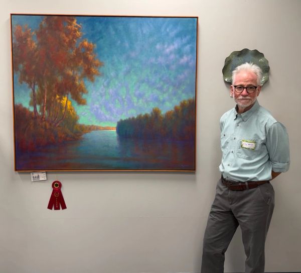 Man posing next to a colorful landscape painting with an award ribbon.