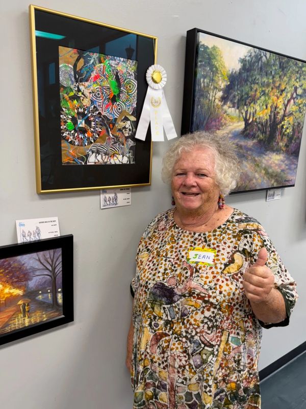 Woman named Jean gives a thumbs up beside her award-winning colorful artwork.