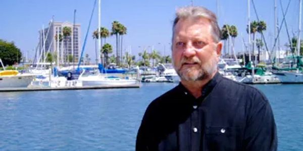 photo of Clarke, black shirt, greying beard, moustache against blue water and sailboats in a marina