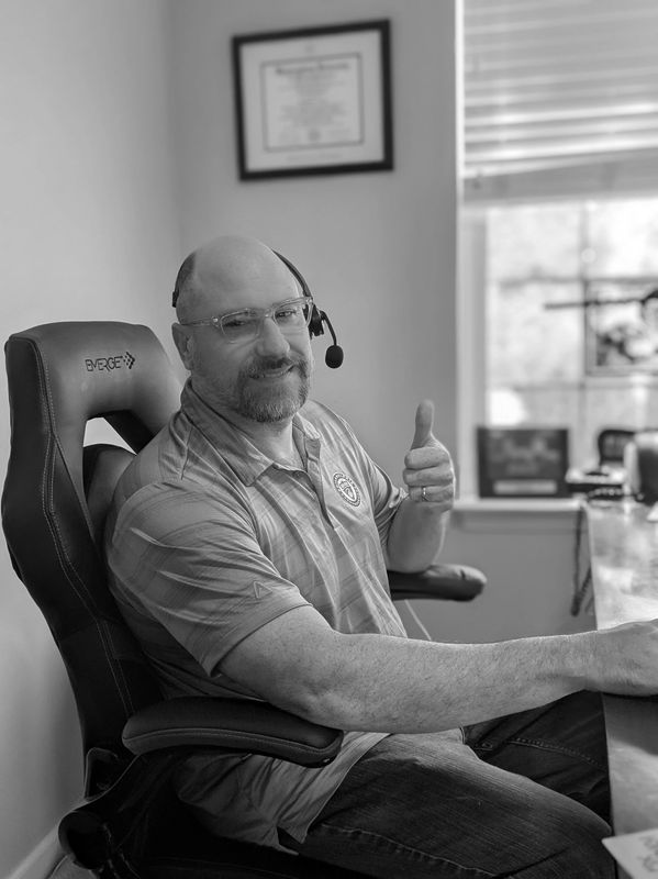 Man in blue polo shirt wearing a headset gives a thumbs up while sitting in an office chair.