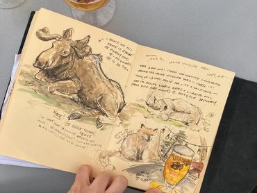Hand holding a sketchbook with animal drawings and a beer glass nearby.