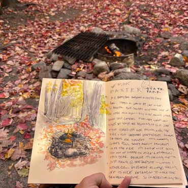 A person sketches a campfire scene in a journal amidst autumn leaves.