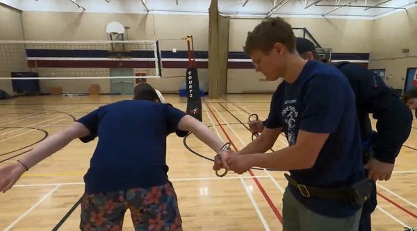 Two men handcuff another man in a gymnasium.