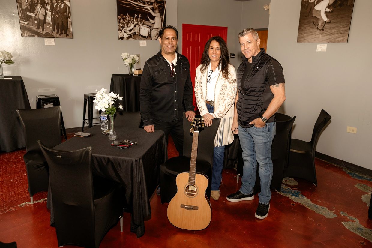 Three people posing indoors with a guitar and black-covered tables.