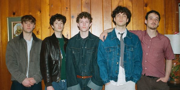 A group of five young men posing indoors against a wooden wall.