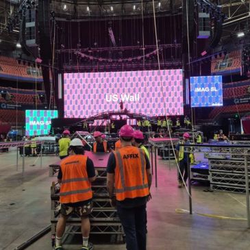 Workers prepare stage lighting and screens for a large indoor event.