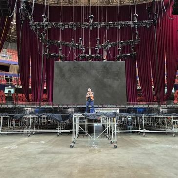 A person stands on a platform in front of a large stage setup with curtains and lighting.