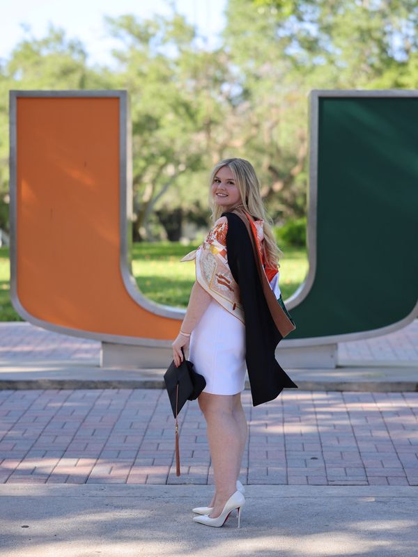 Young woman in white dress holding graduation cap, smiling in front of large U statue.