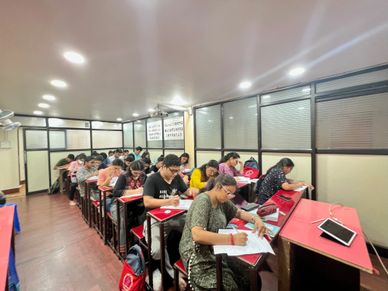 Students focused on writing in a classroom with red desks and bright lighting.