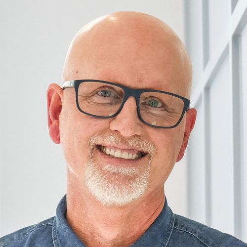 Smiling bald man with glasses wearing a blue denim shirt indoors.