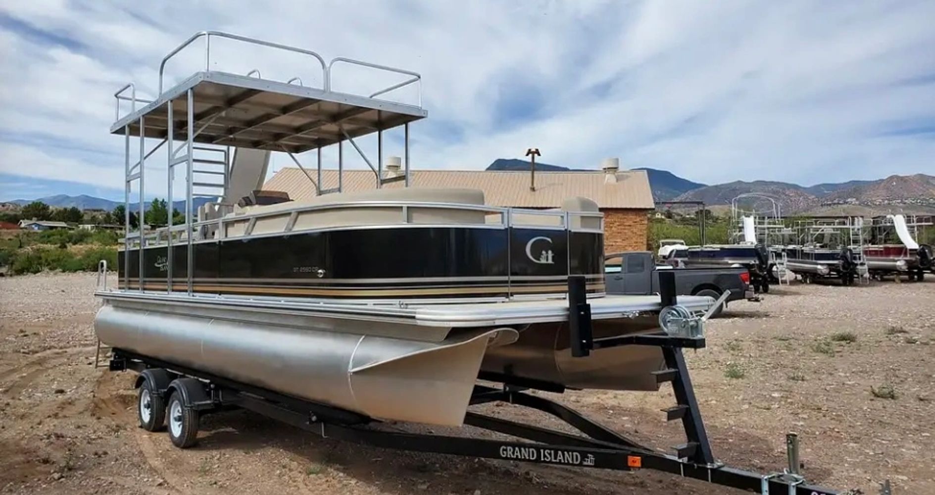 A pontoon boat with an upper deck is parked on a trailer outdoors.